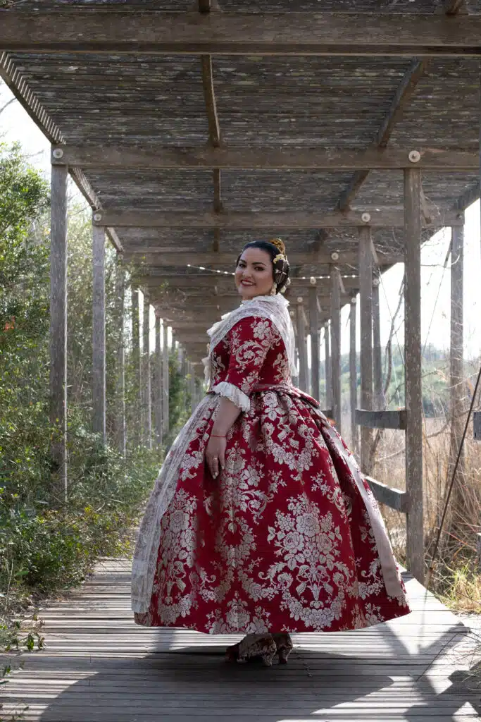 Mujer sonriente con un elegante traje tradicional valenciano rojo y dorado mirando hacia atrás sobre una pasarela de madera bajo una estructura.