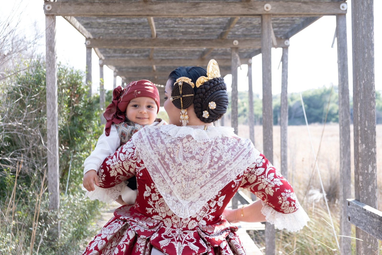 Un instante de ternura capturado bajo la madera: una Fallera con su traje de fallera rojo y dorado sostiene a un niño sonriente con su propio traje fallero, creando una escena encantadora en la pasarela.