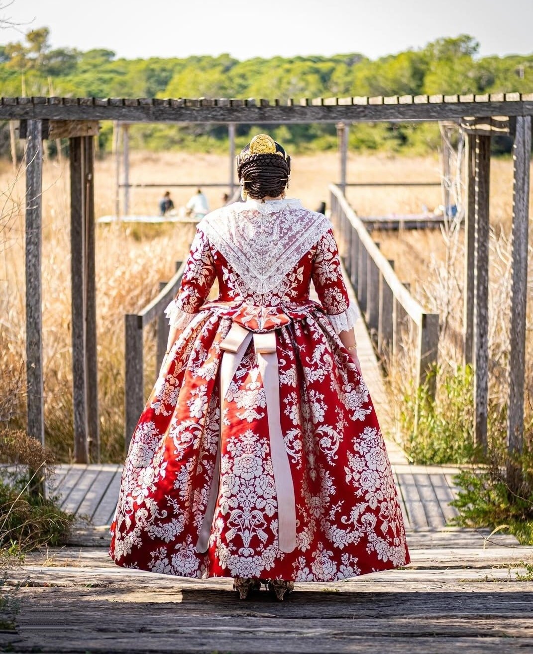 Una vista posterior de una mujer con la vestimenta tradicional valenciana roja y dorada, destacando el elaborado peinado y el lazo trasero, caminando sobre un camino de madera en un entorno natural y soleado.