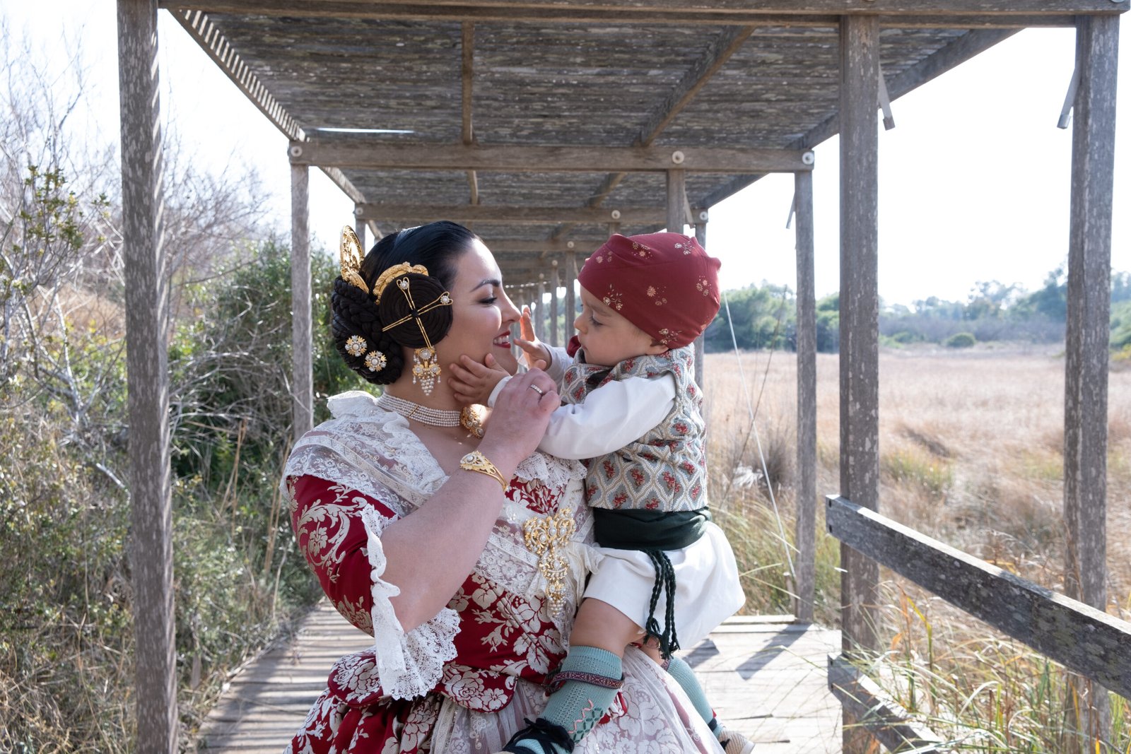 Mujer con traje tradicional valenciano rojo y dorado sosteniendo a un bebé vestido con indumentaria festiva valenciana, bajo una estructura de madera, en la albufera de valencia.