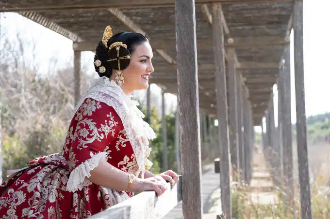 Fallera con un traje regional rojo y dorado, incluyendo un corpiño ajustado y una falda brocada, con la mirada hacia el horizonte bajo un techo de madera.