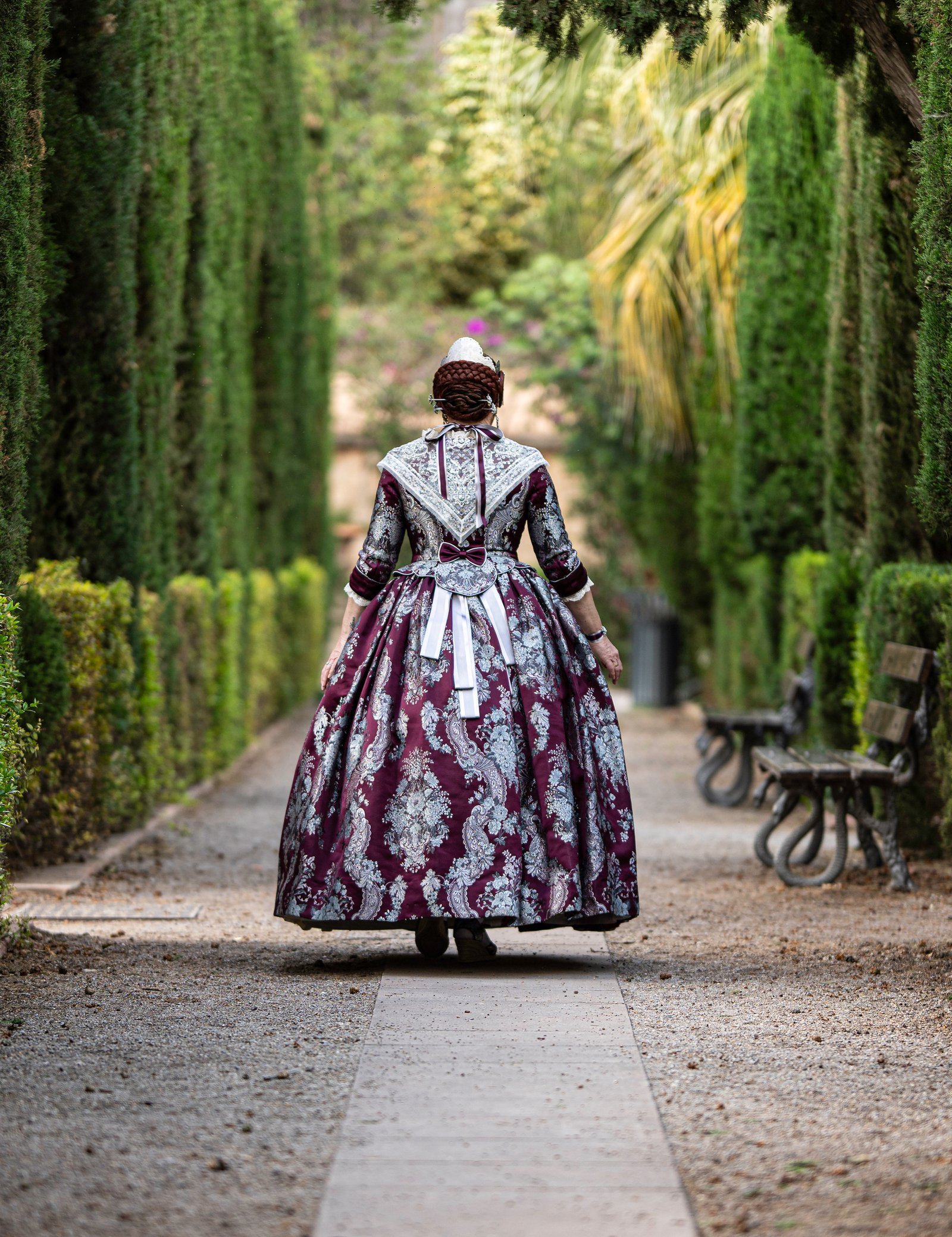 Mujer de espaldas vestida con un traje tradicional valenciano de color burdeos y plata, caminando por un sendero en un jardín con setos verdes y bancos de madera.