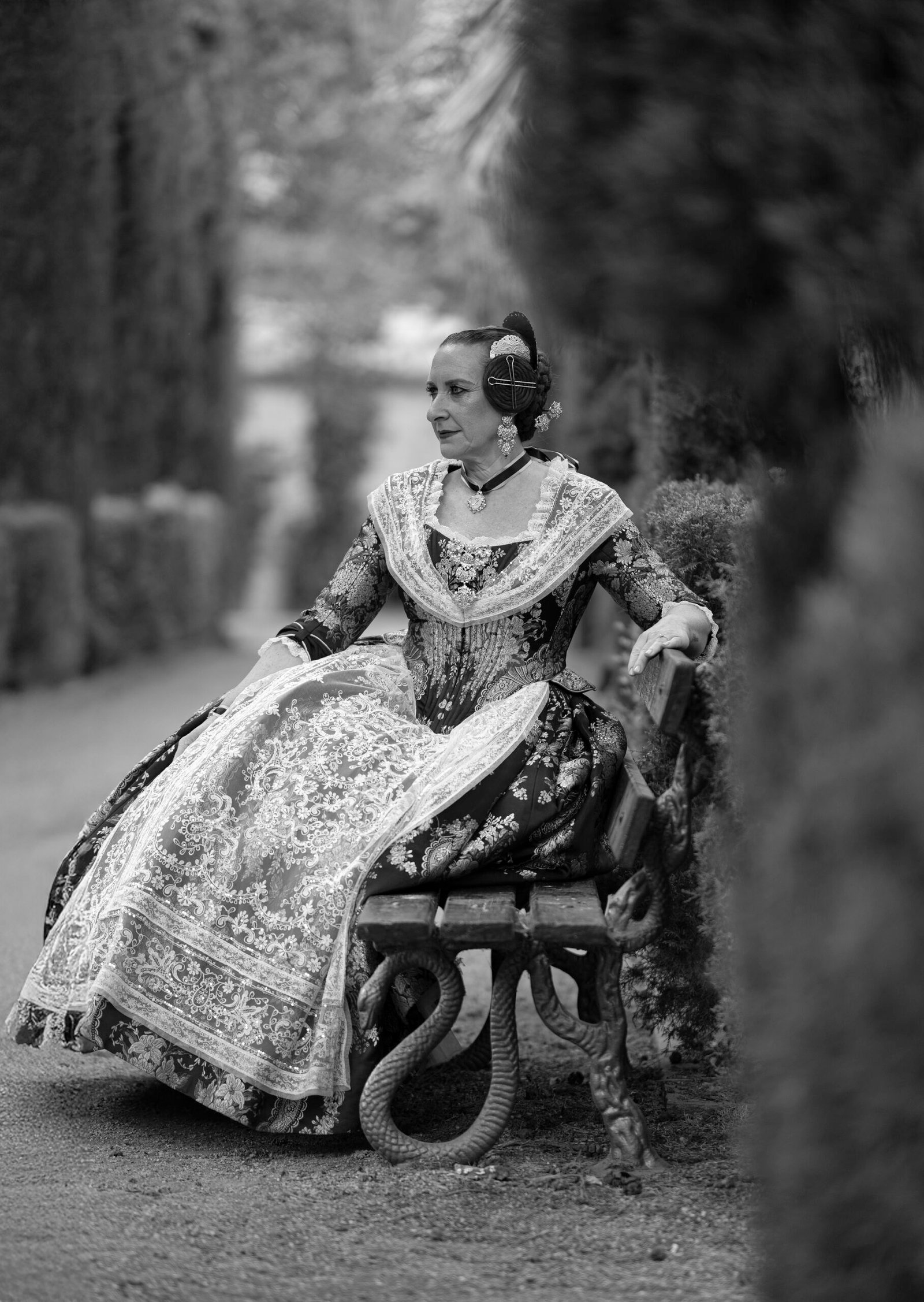 Fotografía en blanco y negro de una mujer elegantemente vestida con un traje tradicional valenciano, sentada pensativamente en un banco de jardín.