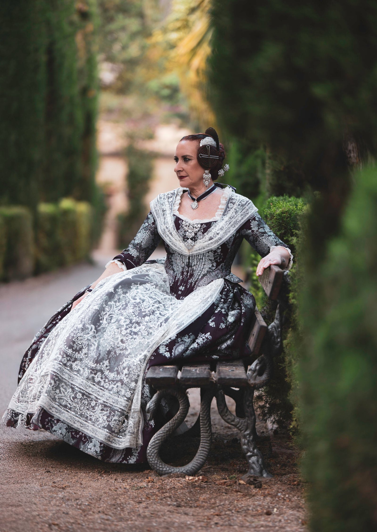 Representación de la belleza de la indumentaria festiva de Valencia a través de una mujer elegantemente vestida con un traje de Fallera en tonos morados y plateados, sentada en un banco de parque, simbolizando la tradición y el folclore de las Fallas.