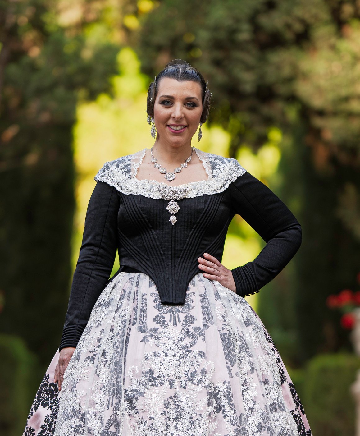 Mujer sonriente con un elegante traje tradicional valenciano negro y blanco, apoyada en una columna de piedra en un entorno ajardinado.