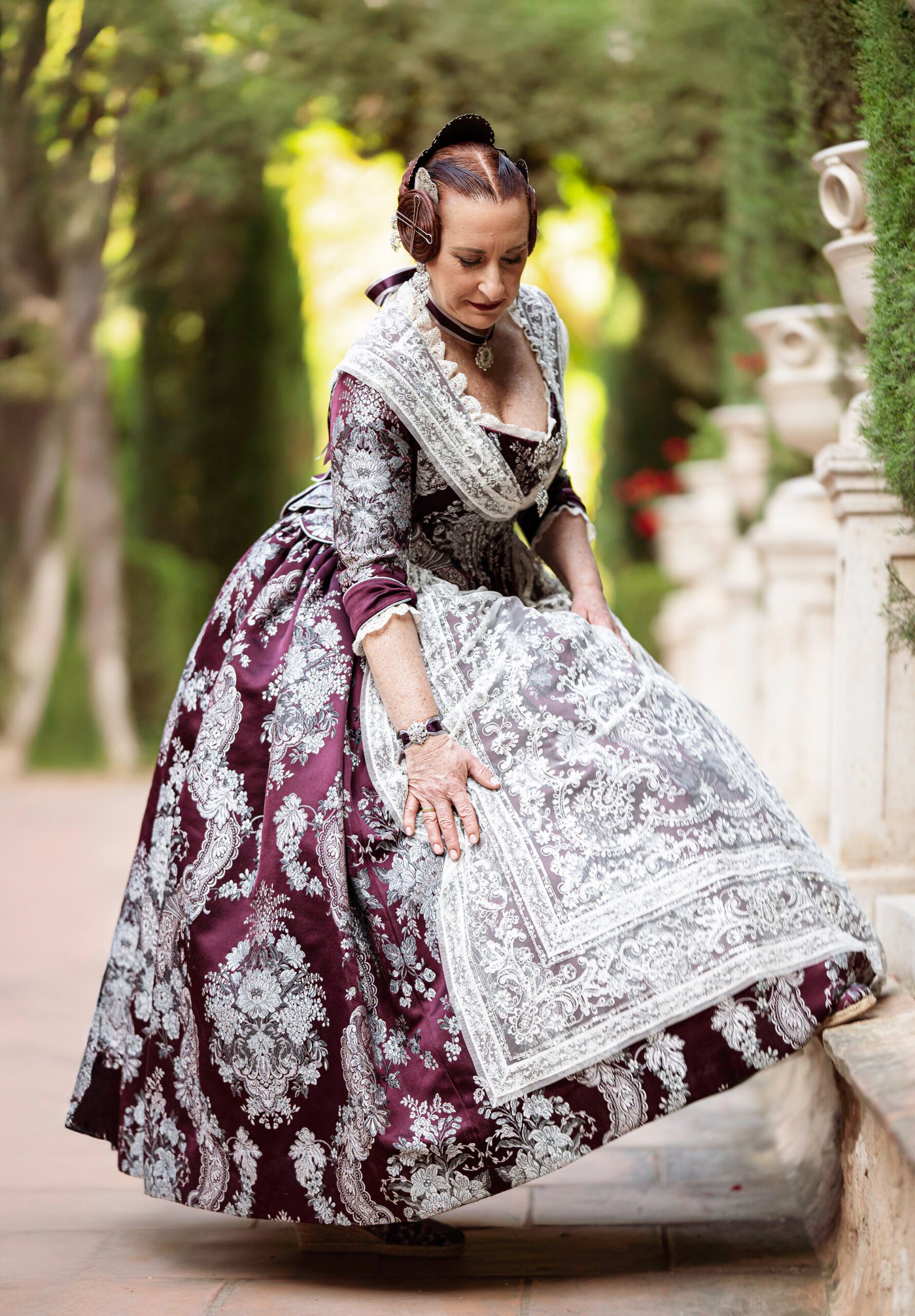 Retrato detallado de Fallera adulta luciendo un hermoso traje de seda brocada en tonos berenjena y plata, destacando la riqueza del encaje y los accesorios, perfecta para 'indumentaria regional valenciana